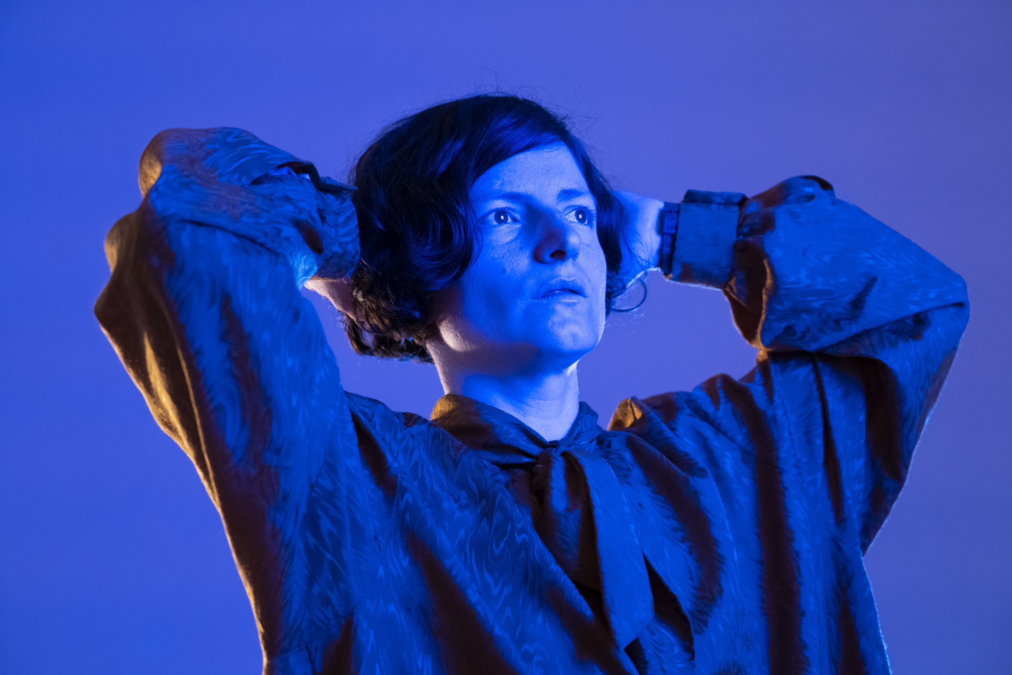 This image shows a close-up of a person surrounded by blue light. The person is holding the hands behind the head and is looking away from the camera.
