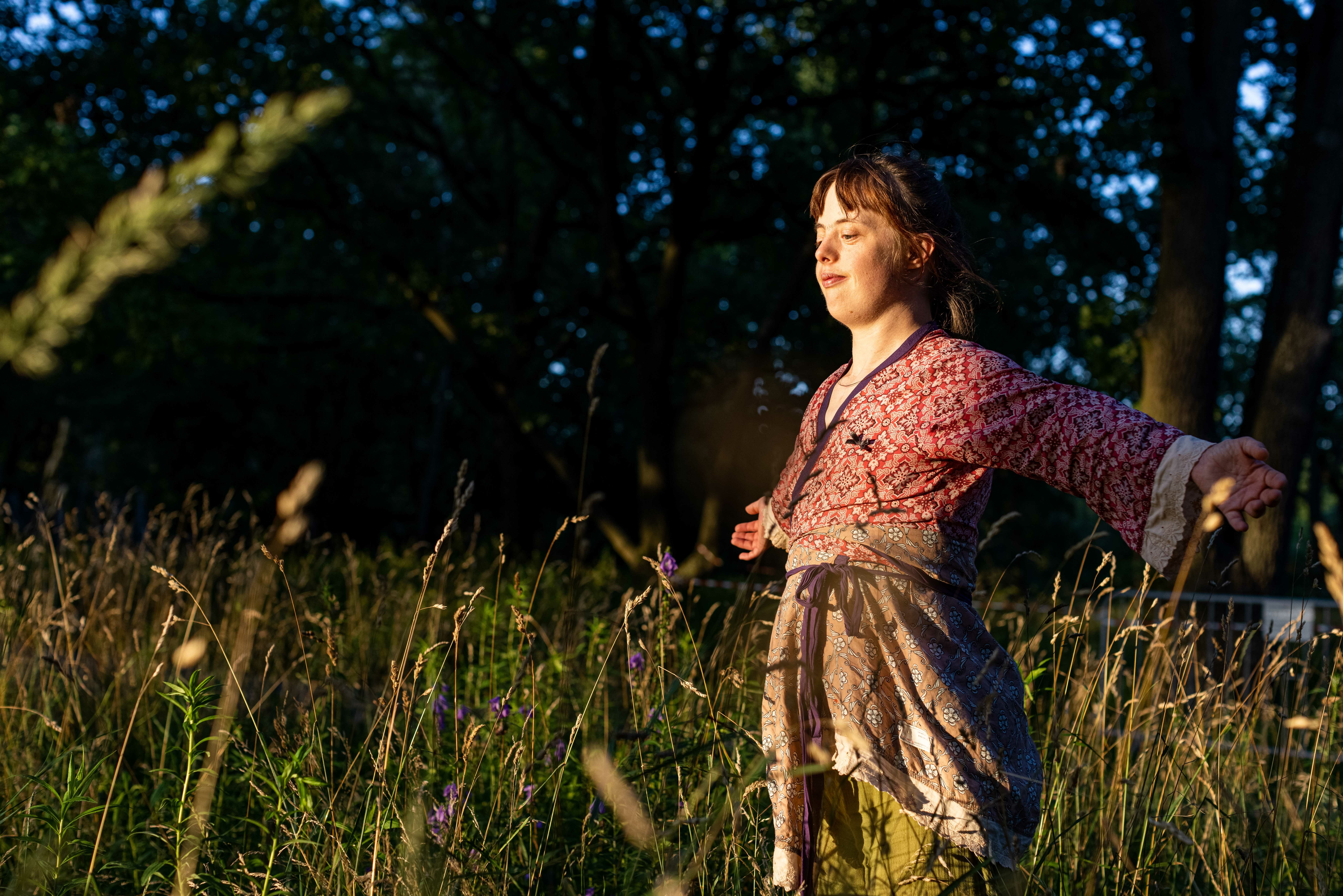 This picture shows a person in red patterned clothing, with her arms spread wide. She is standing in a meadow, with the long grass reaching her hips.