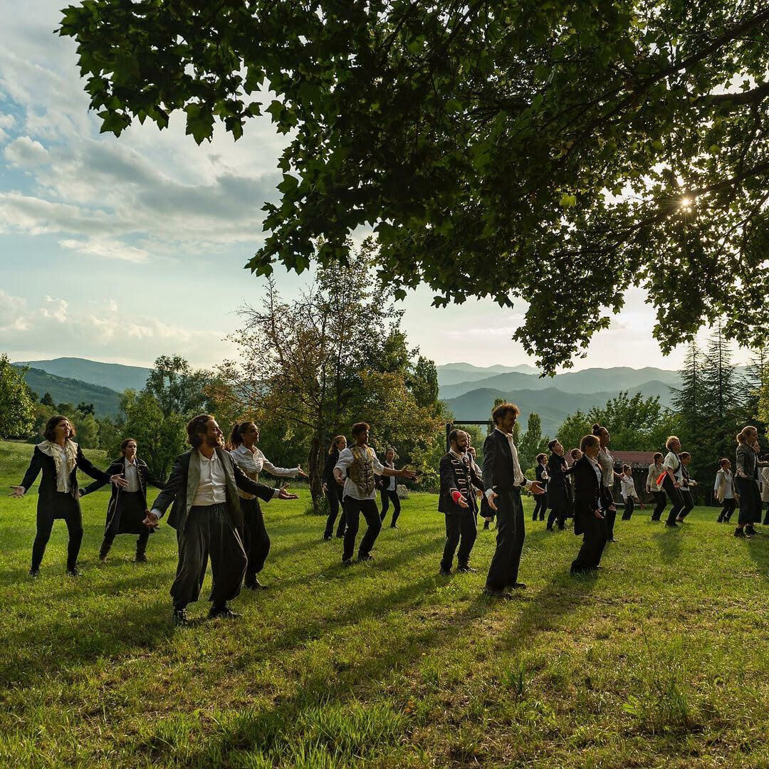 Dieses Bild zeigt eine Gruppe von mehreren Personen. Sie stehen auf einem grasbewachsenen Hang mit einem Wald im Hintergrund. Sie sehen alle sehr unterschiedlich aus. Sie sind alle unterschiedlich gekleidet, und sie machen alle unterschiedliche Gesten.