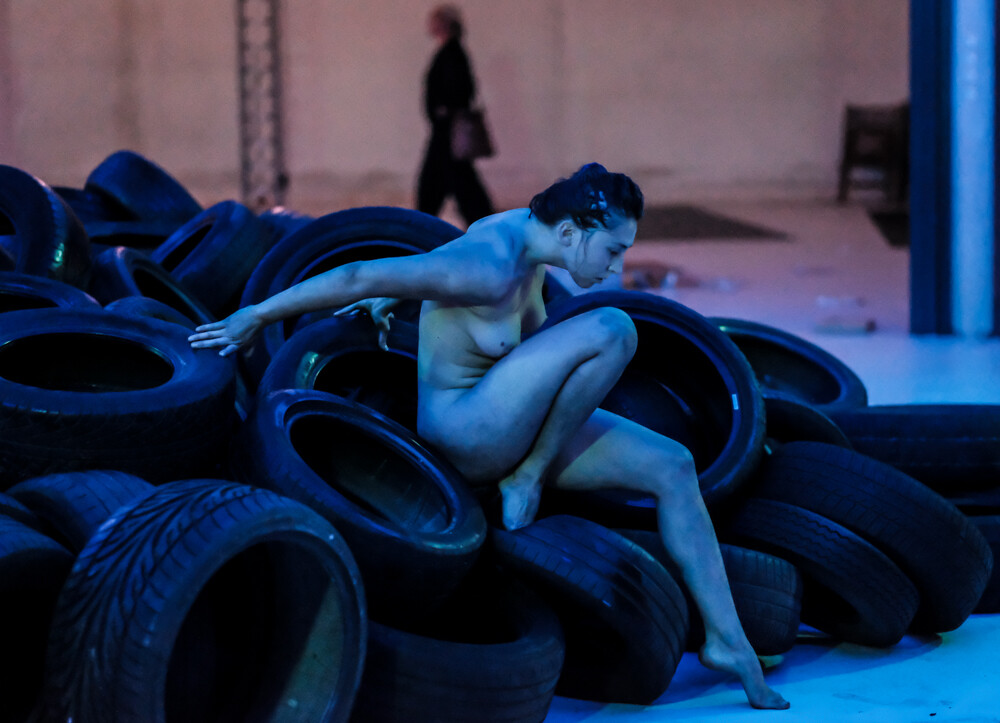 This picture shows a naked woman sitting on a pile of car tyres.