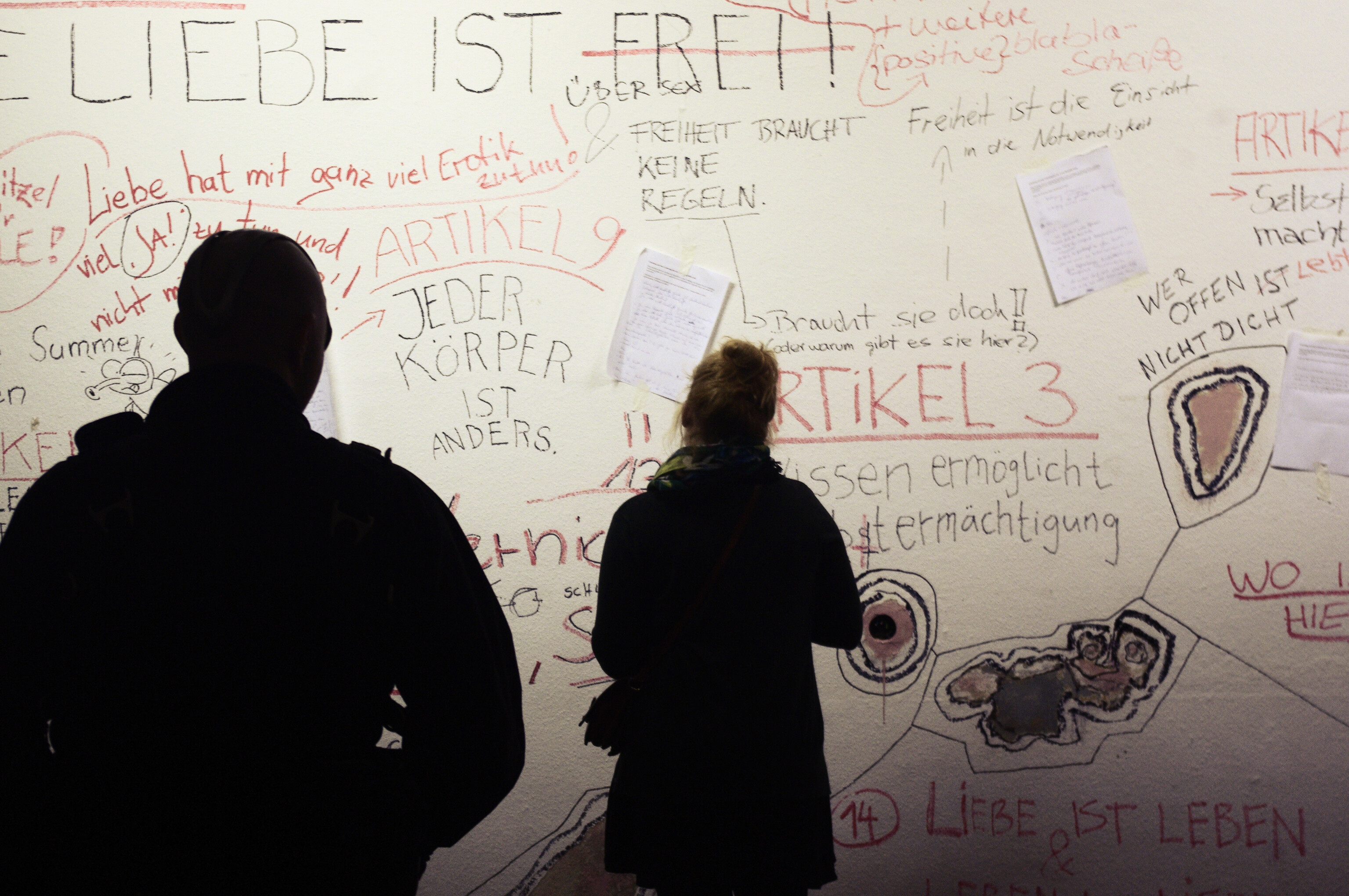 This picture shows two people whose silhouette we see from behind. They are looking at a white wall on which several messages are written in black and red letters.