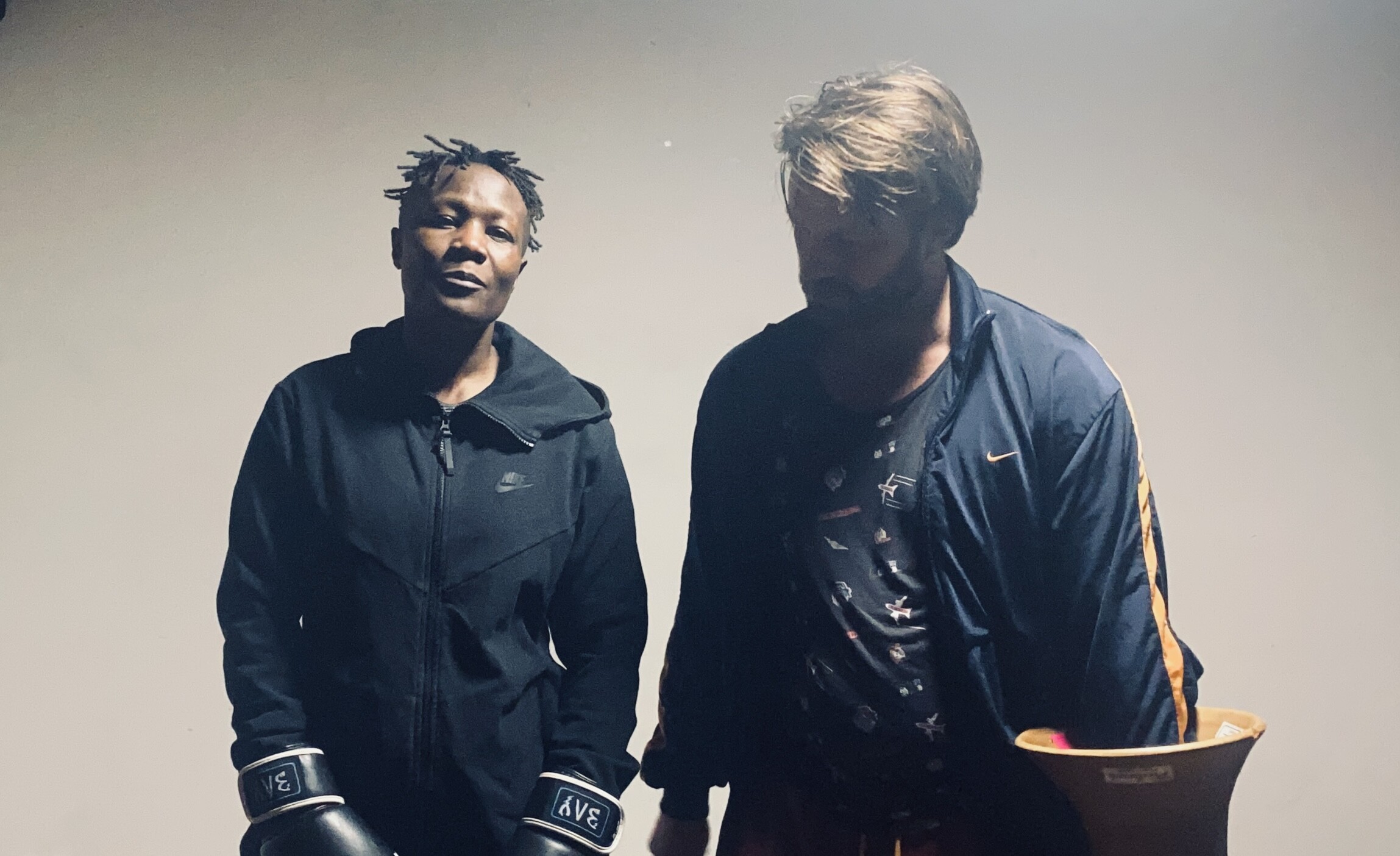 Photo of two people in black sports suits. On the left is a dark-skinned person with black curly hair looking at the camera and on the right is a white person looking at the ground.