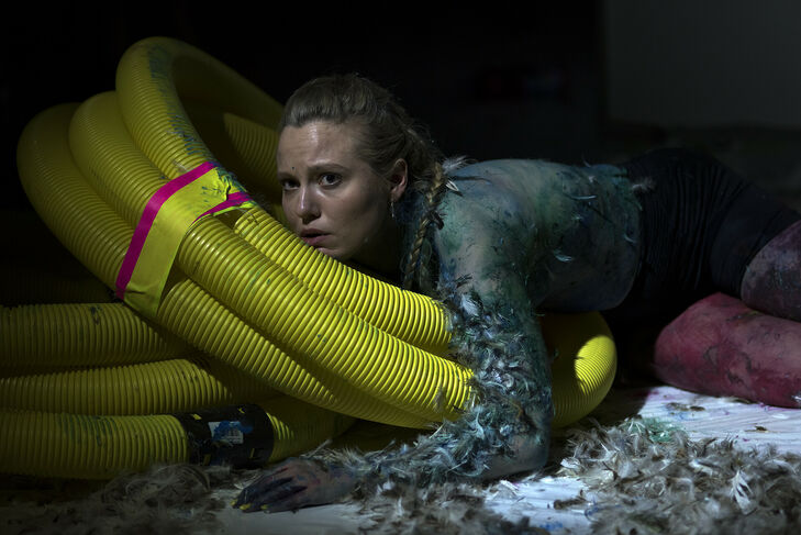 This picture shows a woman lying on the floor. Her upper body is painted with blue and green paint, and she is leaning against yellow plastic pipes. Feathers are stuck to her arm and body. Some feathers are also lying on the floor.