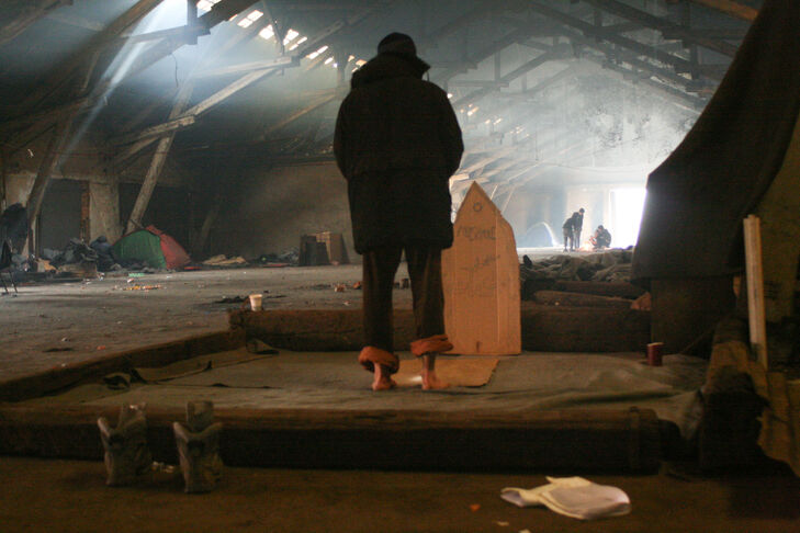 A man stands with his back to the camera in a large abandoned factory building. He is barefoot in an improvised mosque, praying. Several tents can be seen in the factory hall