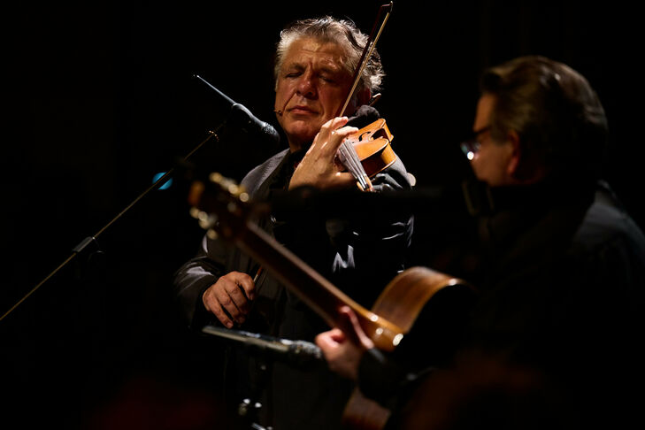 Picture of a concert by Markus Rheinhardt. He is playing his violin with his eyes closed. He is dressed in black. A guitarist is playing next to him.