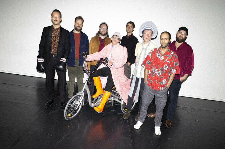 Bild von der inklusiven Band namens Station 17, bestehend aus acht Männern. Links im Bild steht ein Künstler mit Handschuhe und mittig sitzt ein Mann auf einem Dreirad.