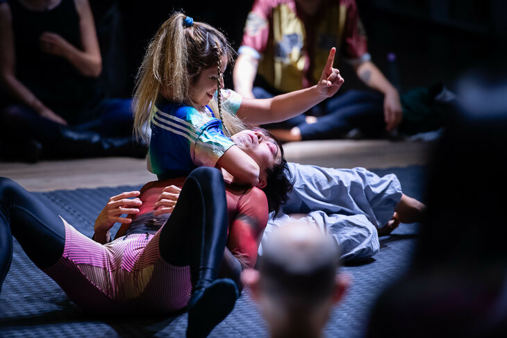 Photo of two women wrestling on the floor, one woman wraps her arm around the neck of the other woman and points her finger upwards with her other hand.