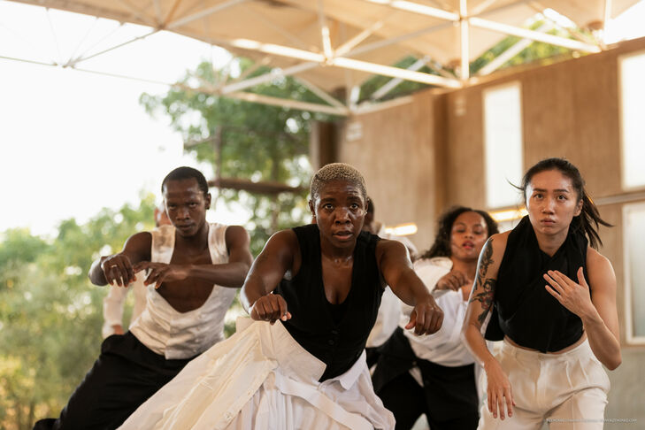 Group of people dancing. They wear white and black clothing and are in a large, open space for training.