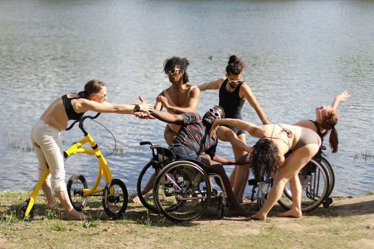 Photo by a lake of six people pulling each other by the arms. Two are in a wheelchair and they are all wearing white sunglasses.